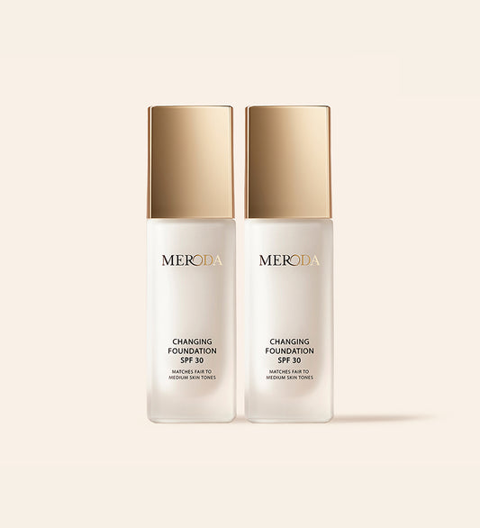 Zwei Flaschen Changing Foundation SPF 30 mit goldenen Kappen nebeneinander auf einem cremefarbenen Hintergrund.